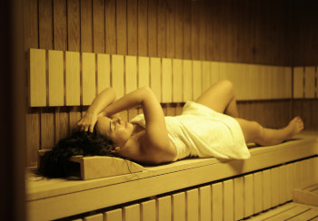 Frau mit dunklen Haaren liegt auf einer Sitzbank in einer Sauna. Sie ist mit einem weißen Handtuch bedeckt.