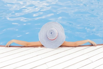 Eine Frau entspannt am Beckenrand Ihres Pools. Sie trägt einen Hut
