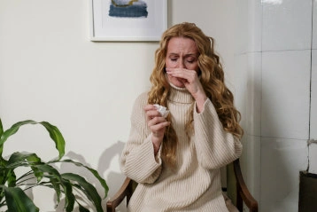 Erkältete Frau hält sich die Nase vor das Gesicht. In der anderen Hand hält sie ein Taschentuch